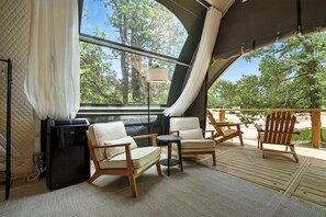 Ein gemütlicher Sitzbereich im Freien mit zwei Stühlen und einem kleinen Tisch, einem Deckenventilator und Blick auf Bäume durch ein großes Fenster.