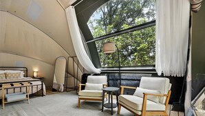 Une pièce aménagée comme une tente, avec un lit, deux chaises, une petite table et une vue sur les arbres à travers de grandes fenêtres.