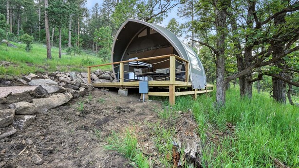 A dome-shaped cabin with a wooden deck and a table and chairs inside, surrounded by a forested area. - Glamping in Nature Near Mt. Rushmore (Hermosa)