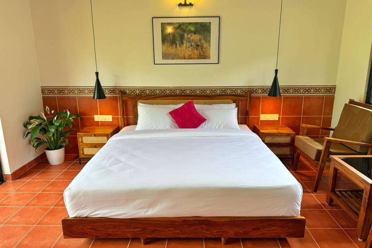 Een net opgemaakt bed met witte lakens en een rode kussen, zijkanten begrensd door twee nachtkastjes met lampen, in een kamer met terracotta tegels en een decoratief wandpaneel.