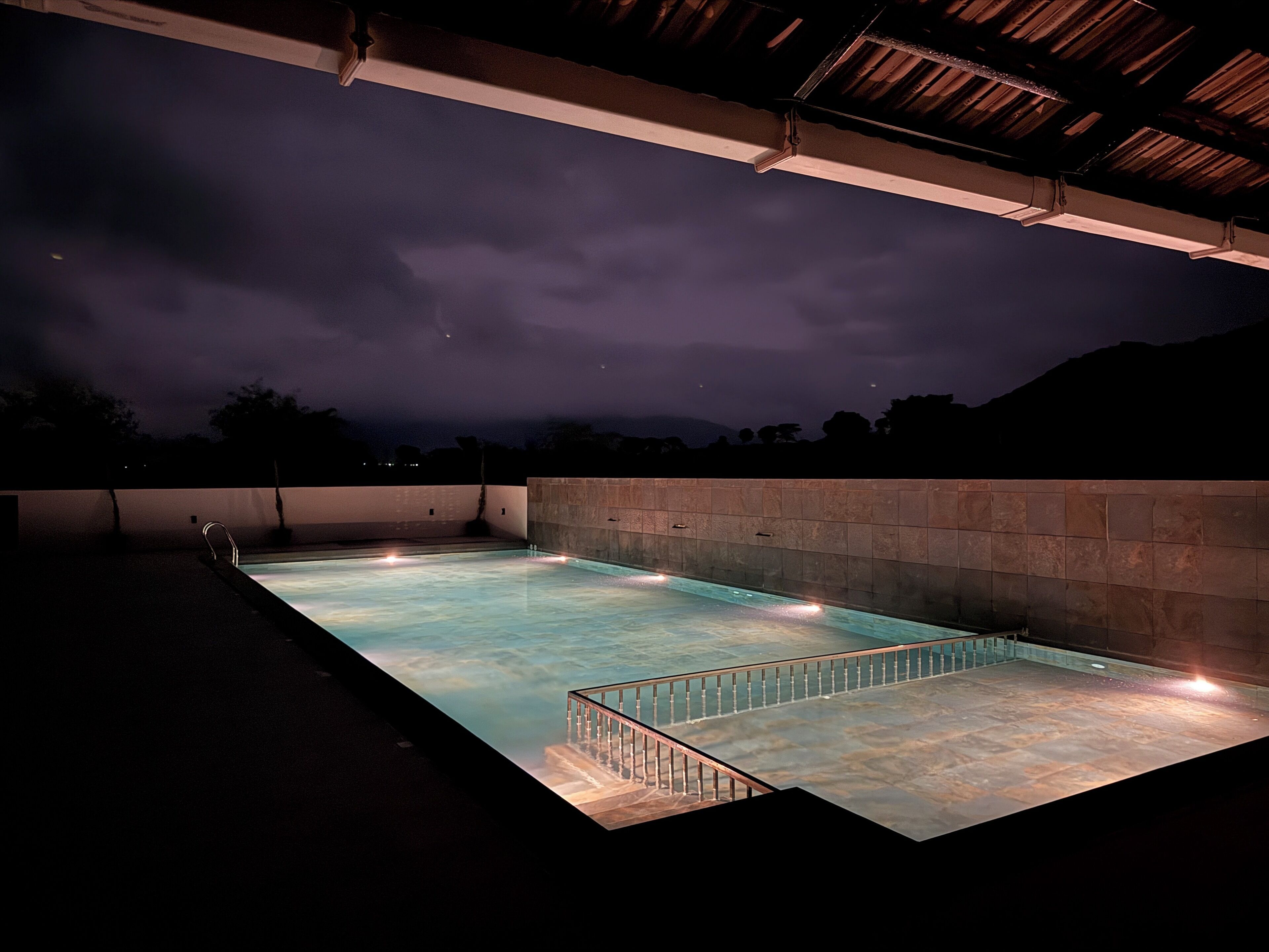 Una piscina al aire libre con pared de piedra y barandilla de metal, bajo una zona cubierta con techo inclinado.