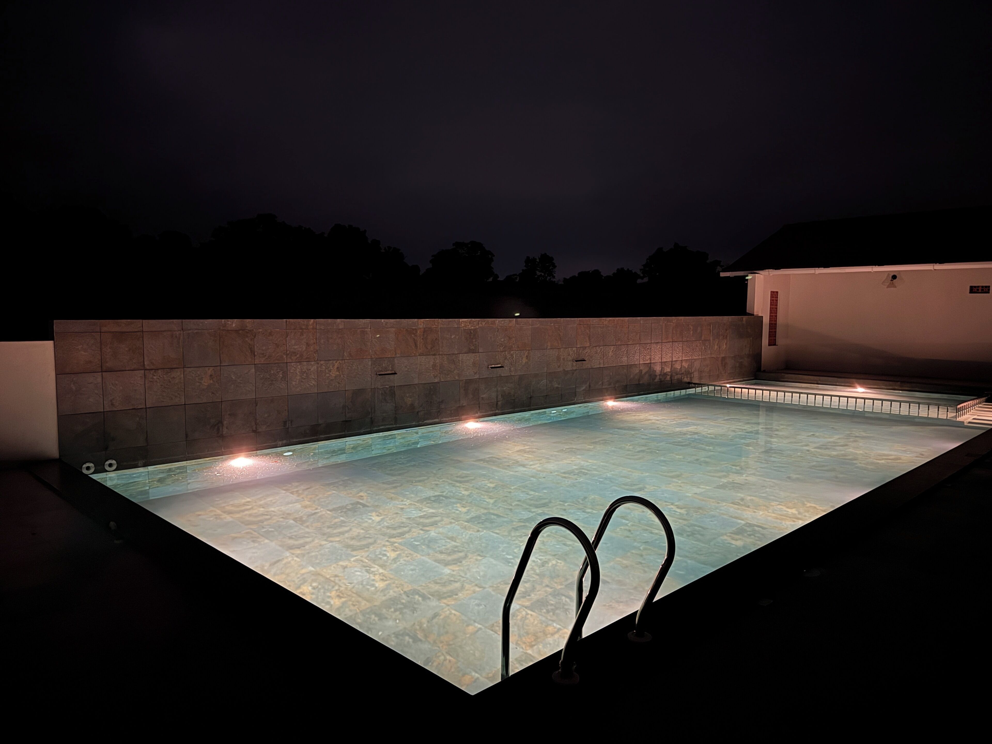 A rectangular swimming pool with a stone wall and metal railings, surrounded by a dark night sky.