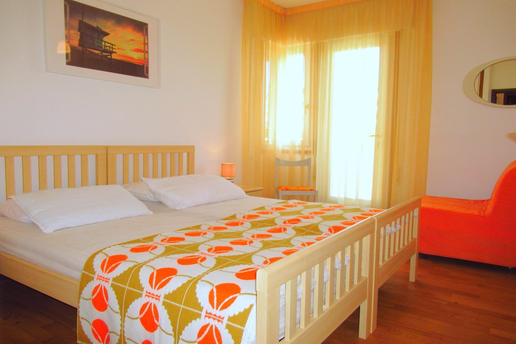 A bedroom with a wooden bed, a nightstand, a chair, a window with curtains, and a wall-mounted mirror.