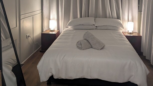 Un lit bien fait, avec une couette blanche et deux oreillers, une lampe de chevet et une serviette pliée posée sur le lit.