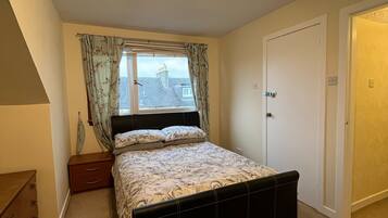 Un dormitorio con cama, mesita de noche, cómoda, ventana con cortinas y puerta.