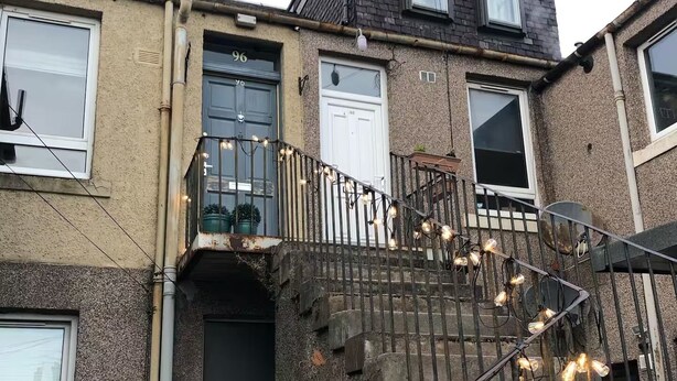 A multi-story residential building with a visible staircase, a small wooden structure at the entrance, and a balcony with string lights.