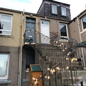 Un edificio residenziale a più piani con una scala visibile, una piccola struttura in legno all'ingresso e un balcone con luci decorative.