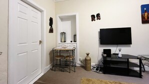 A living room with a white door, a TV mounted on the wall, a small table with chairs, and a decorative vase.