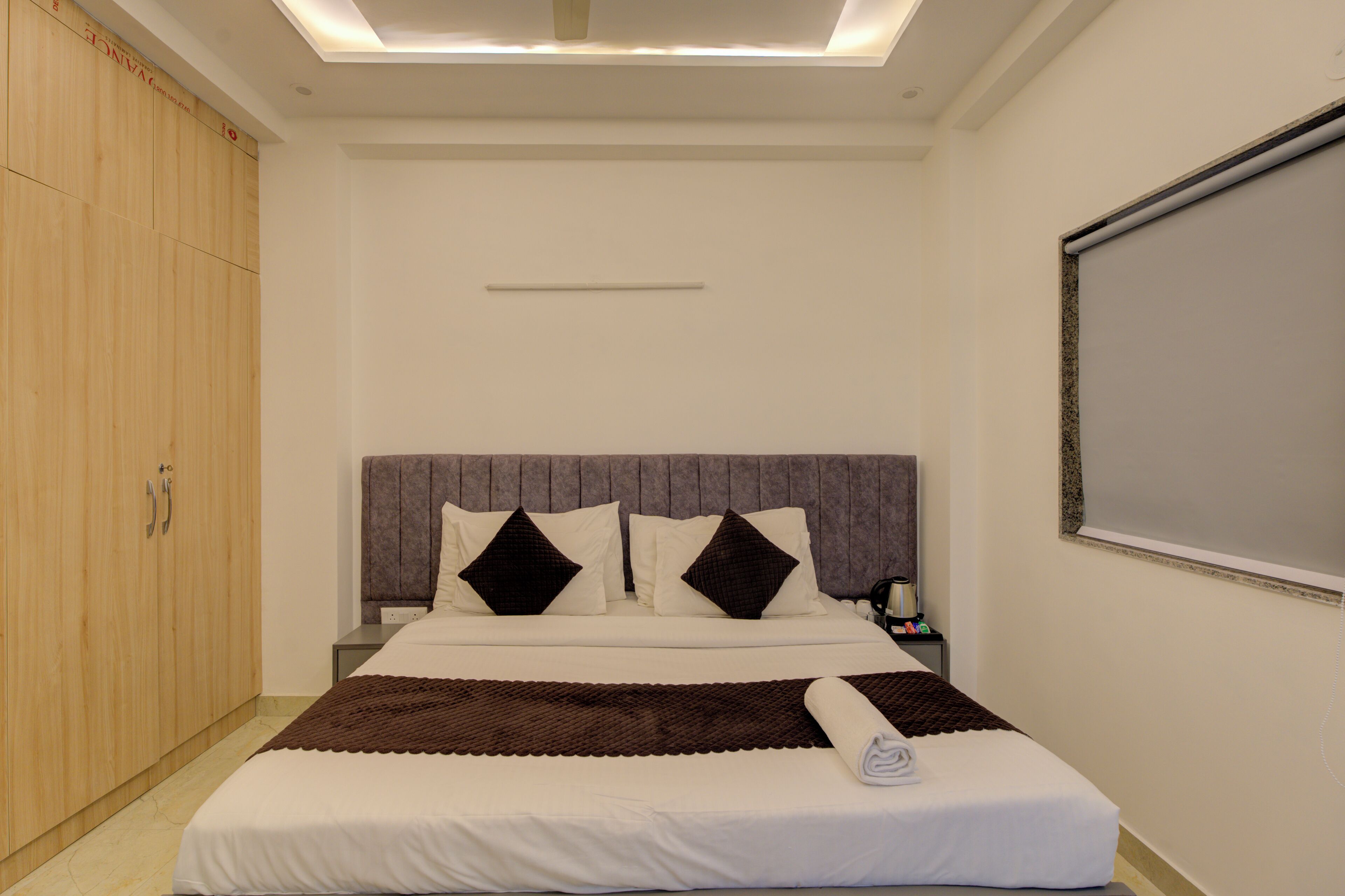 Ein ordentlich bezogenes Bett mit einer braunen Tagesdecke und einem aufgerollten Handtuch, ein Holzschrank und ein großes Fenster mit Blick zum Himmel.
