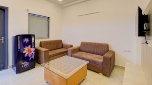 A living room with two brown sofas, a glass coffee table, a refrigerator with floral design, a ceiling fan, and a mounted TV.