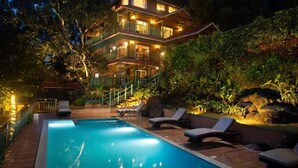 A multi-story building with a swimming pool, lounge chairs, and a well-lit exterior. - Bamboo Dale Resort (Udumbanchola)