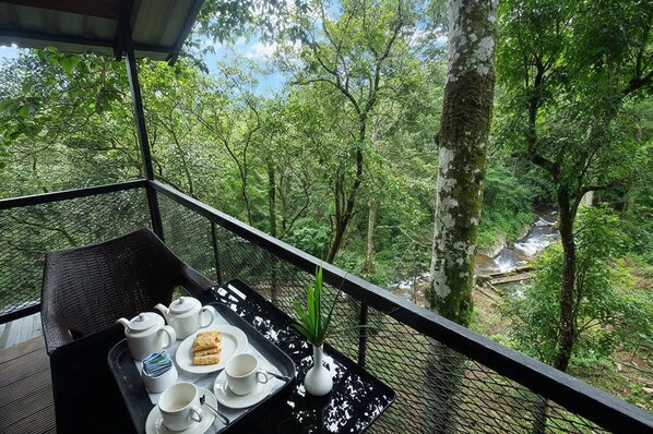 Un balcon avec une table dressée pour le thé et les biscuits, donnant sur une zone boisée traversée par un ruisseau.