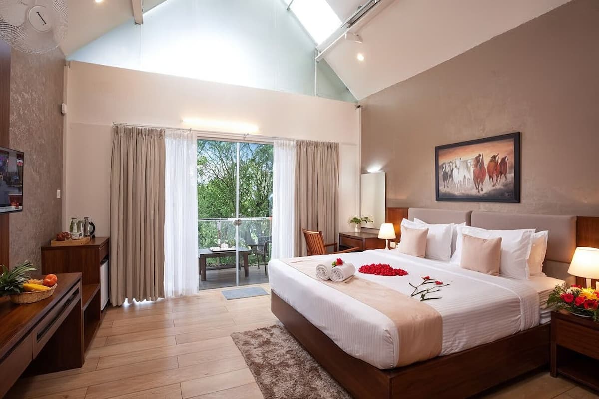 Un dormitorio amplio con una cama grande, un balcón con cortinas, un cuadro en la pared y una mesita de noche de madera con una lámpara.