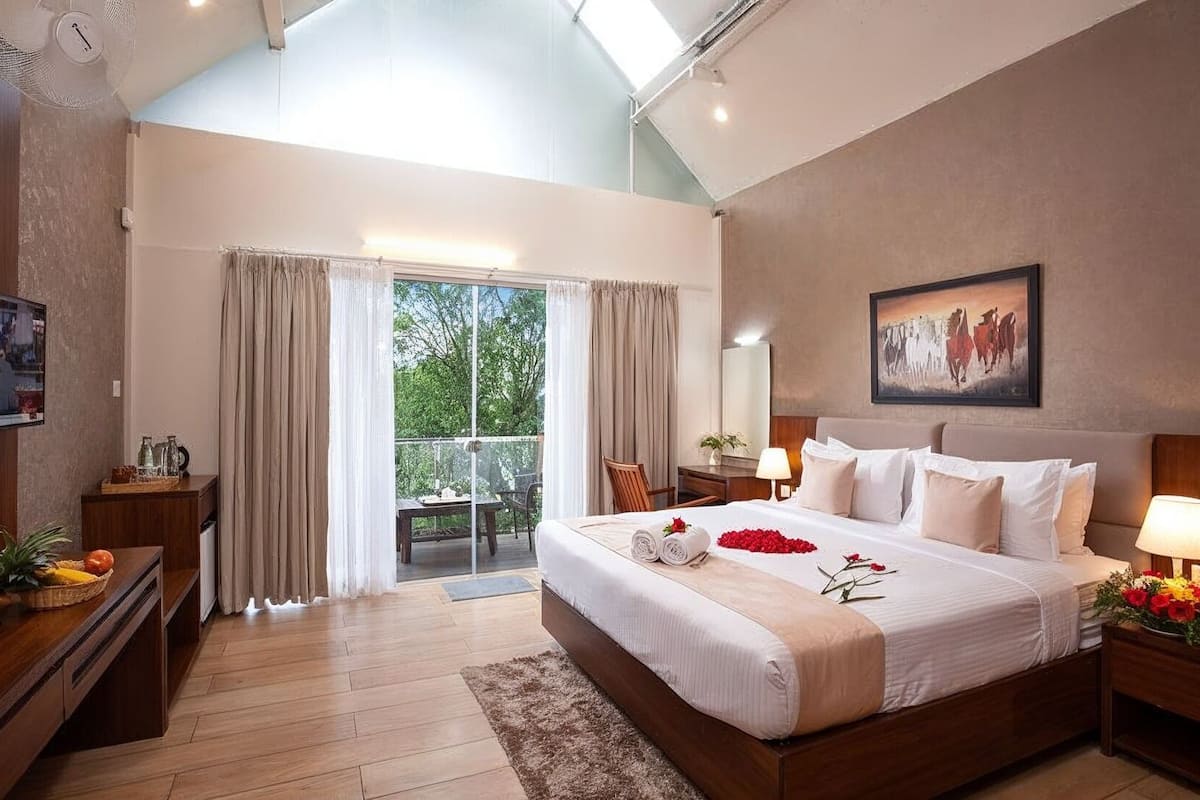 Un dormitorio amplio con una cama grande, un balcón con cortinas, un cuadro en la pared y una mesita de noche de madera con una lámpara.