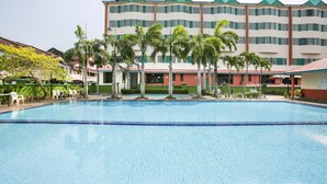 Una piscina con borde de baldosas, rodeada de palmeras y un edificio de varios pisos con techos verdes y paredes blancas.