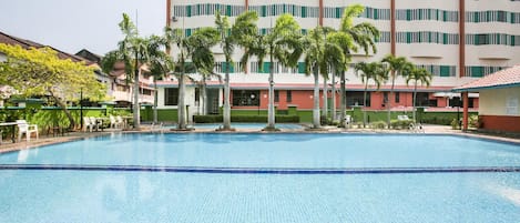 A swimming pool with a tiled deck, surrounded by palm trees and a multi-story building with green roofs and white walls.