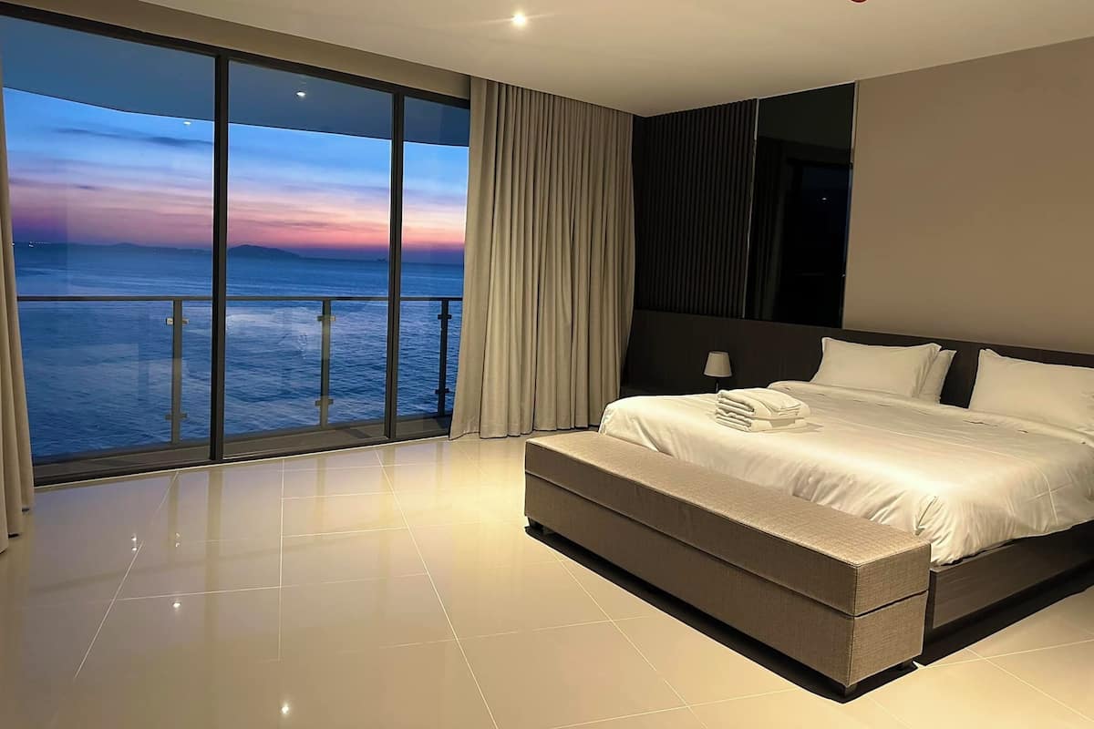 Une chambre d’hôtel moderne dotée d’un grand balcon, d’un lit avec du linge de lit blanc et offrant une vue sur l’océan au coucher du soleil.