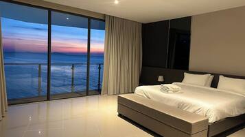A modern hotel room with a large balcony, a bed with white linens, and a view of the ocean at sunset.