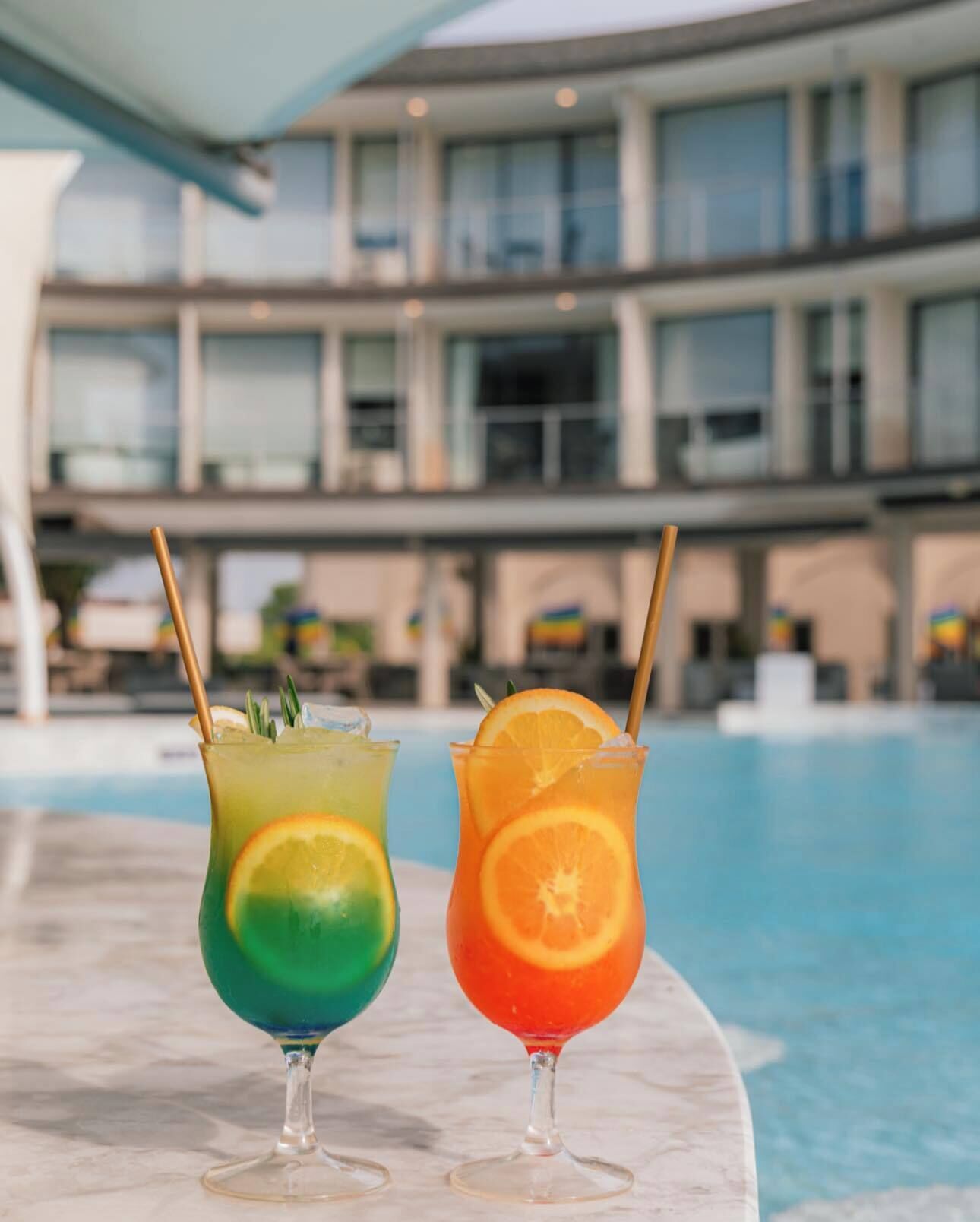 Two cocktails with straws and garnishes on a poolside table.
