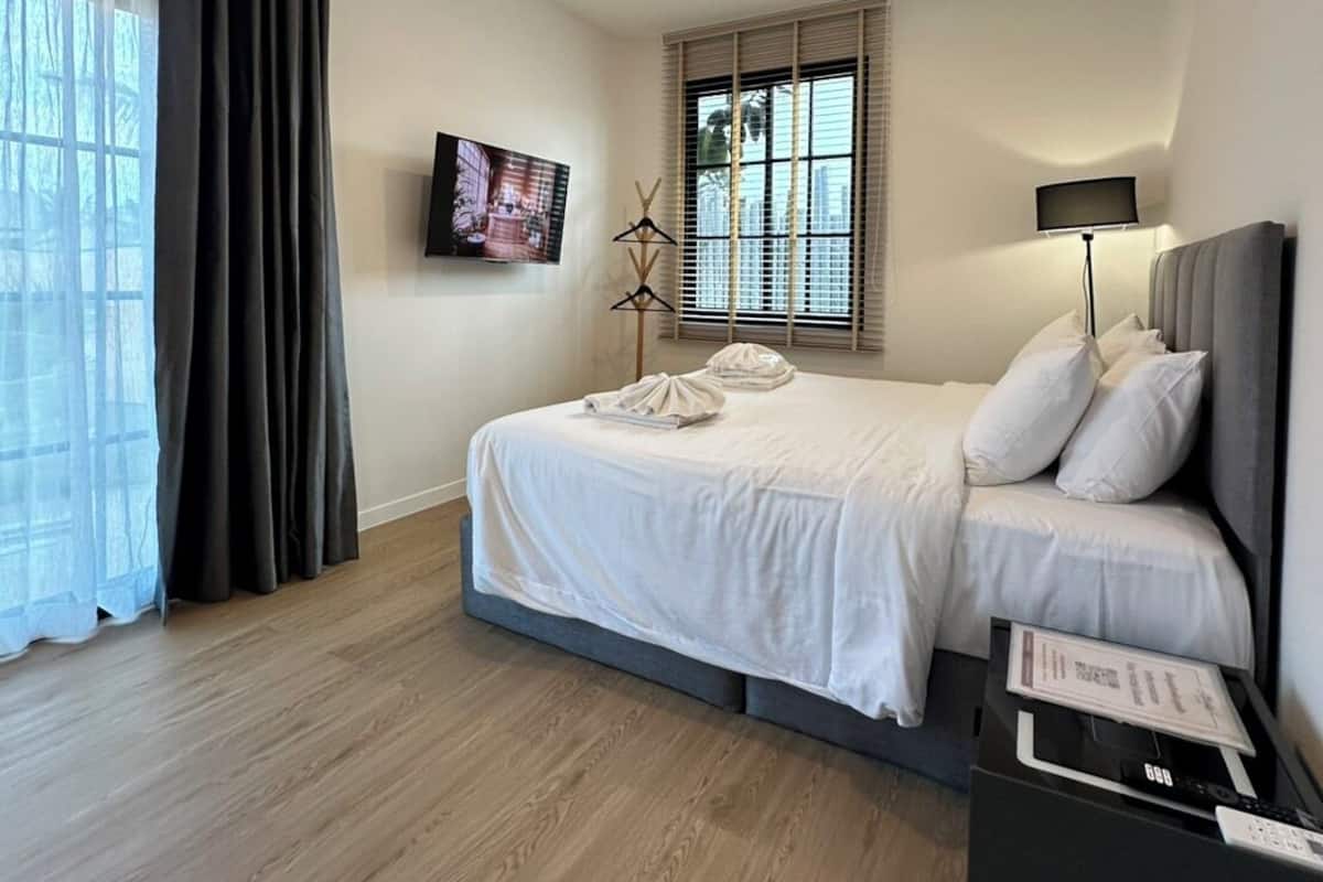Une chambre d’hôtel moderne avec un grand lit, un téléviseur fixé au mur, une table de chevet avec une lampe et une vue sur la ville par la fenêtre.