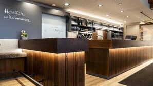 A modern reception area with a wooden reception desk, a seating area, and a bar with shelves of bottles and glasses. - Eisenbocks Strasser Hof (Straß im Straßertale)