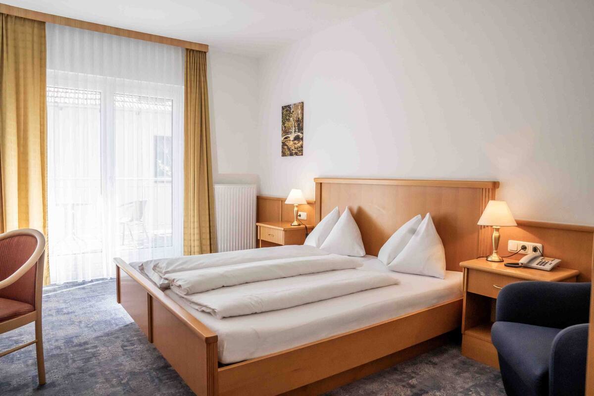 Habitació d'hotel amb un llit gran, dues taules de nit amb làmpades, una cadira i una finestra amb cortines.