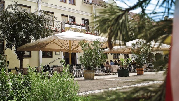 An outdoor cafe with tables and chairs, surrounded by potted plants and a building with a sign. - Eisenbocks Strasser Hof (Straß im Straßertale)
