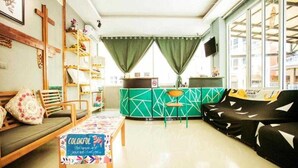 Un salón acogedor con un sofá, un banco de madera y una mesa de colores. Hay una cortina verde y una ventana con vistas a edificios.