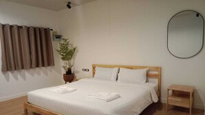 Una cama bien hecha con sábanas y toallas blancas, una pequeña mesita de noche de madera, una planta en maceta y un espejo fijado a la pared.
