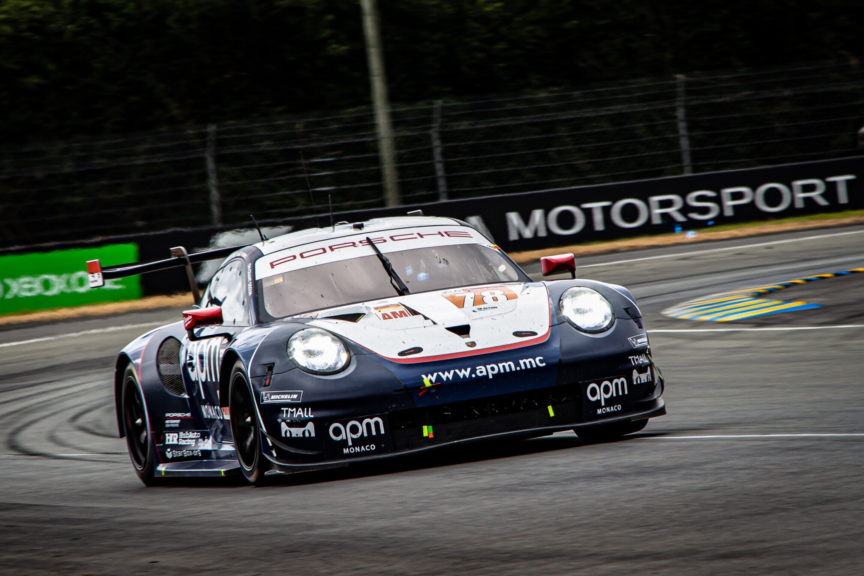 Un auto de carreras en una pista, con logotipos de patrocinadores visibles y un letrero que dice 'MOTORSPORT' al fondo.