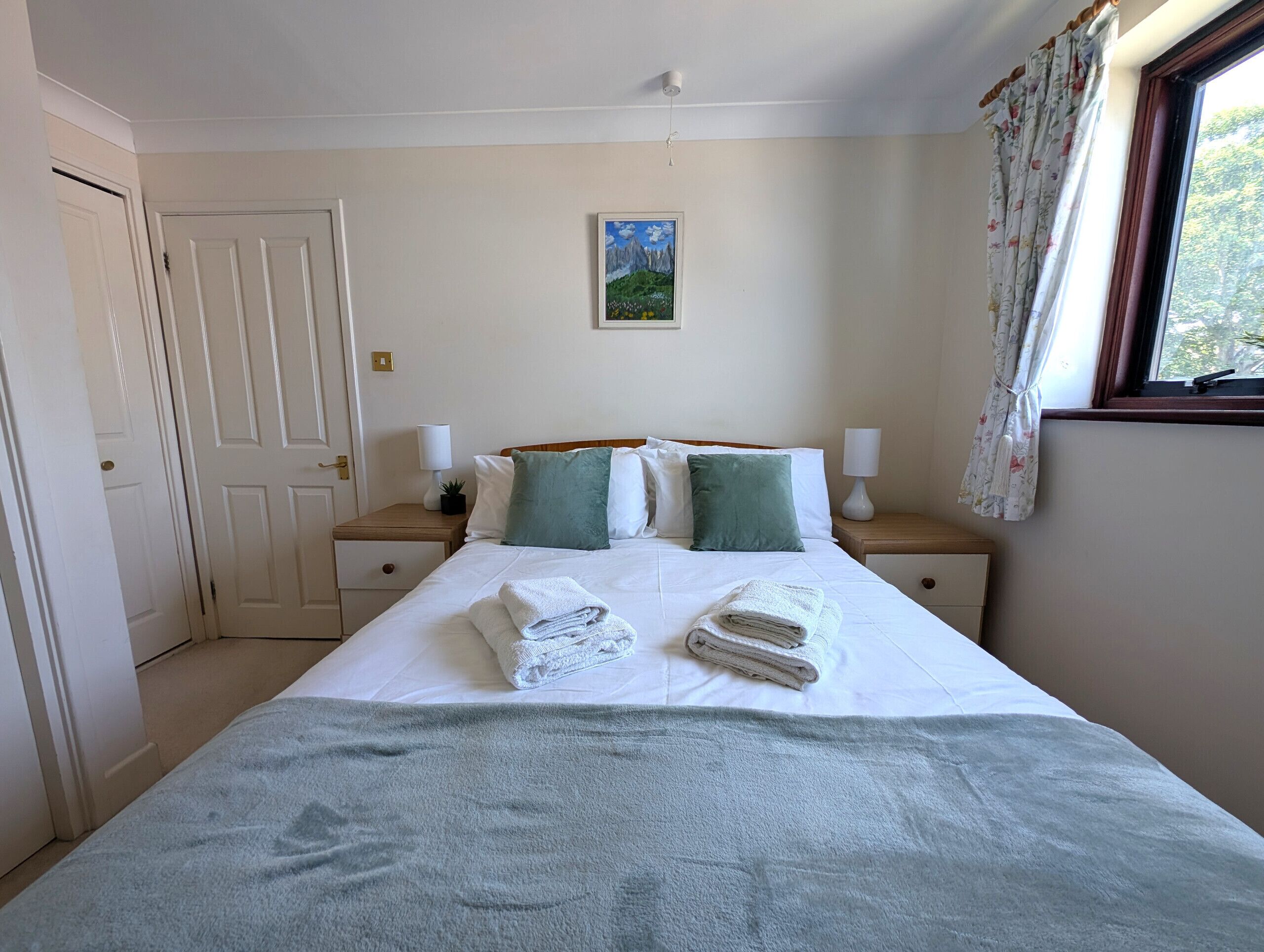 A neatly made bed with folded towels, a bedside table with a lamp, and a window with curtains.