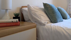 A neatly made bed with white and teal pillows, a bedside lamp, and a stack of books on a wooden nightstand. - 19 Seaview Bay-Isle of Wight (Seaview)