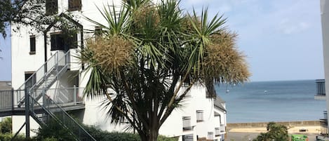 A white building with a staircase and balconies, a palm tree in front, and a view of the sea.
