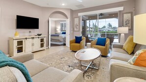 A spacious living room with a flat-screen TV, a sofa set, a coffee table, and a view of a balcony with a pool.