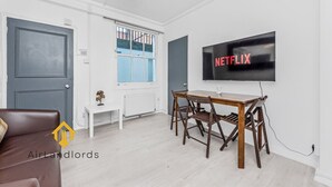 A modern living room with a TV displaying 'NETFLIX', a wooden dining table, a brown leather sofa, and a window with blue shutters. - Exclusive 4 bedroom apartment in Russell Square (Greater London)