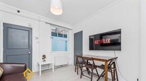 Een moderne woonkamer met een televisie waarop 'NETFLIX' staat, een houten eettafel, een bruine leren bank en een raam met blauwe luiken.