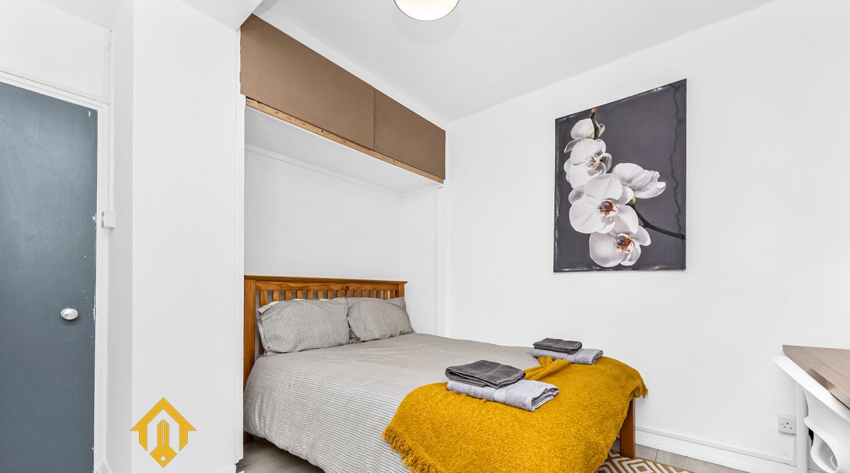 Une chambre moderne avec un lit, une tête de lit en bois, un plaid de couleur moutarde et un tableau représentant une orchidée, encadré et accroché au mur.