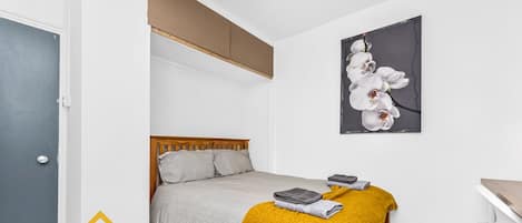 Une chambre moderne avec un lit, une tête de lit en bois, un plaid de couleur moutarde et un tableau représentant une orchidée, encadré et accroché au mur.