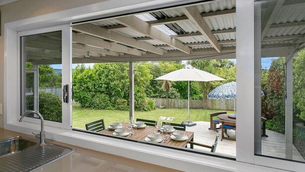 Eine moderne Küche mit einem großen Schiebeglasfenster, das zu einem Essbereich im Freien führt. Dort steht ein Holztisch für vier Personen, ein großer Sonnenschirm und ein Blick auf einen üppigen Garten.