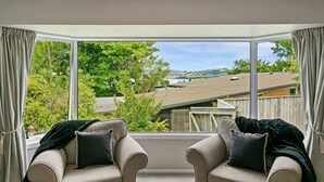 Ein Zimmer mit großem Fenster und Blick auf eine Wohngegend mit Bäumen und Häusern. Zwei gepolsterte Sessel mit Kissen sind vorhanden.