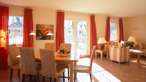 A dining area with wicker chairs, a wooden table, and a bowl of fruit. There are two windows with red curtains, and a painting on the wall. - Kustenwaldvilla in Dierhagen (Ostseebad Dierhagen)