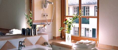 A bathroom with a sink, a hairdryer, a window with a view of buildings, and a vase with flowers.
