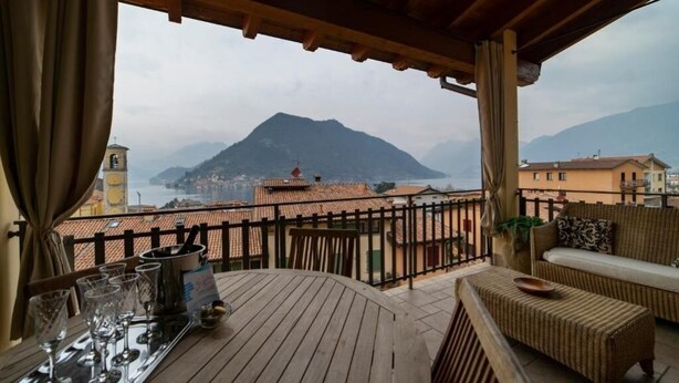 A wooden deck with a table set for dining, a wicker sofa, and a view of a mountainous landscape with a body of water in the background. - ROMANTIC LAKE VIEW HOUSE (Sulzano)