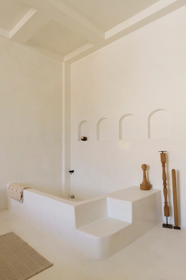 A minimalist bathroom with a bathtub, a wooden bench, and a small rug. - The Hidden Home (Patong)