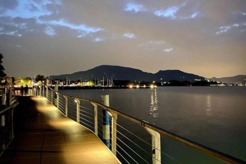 A waterfront promenade at dusk with a calm river, distant mountains, and a well-lit walkway.