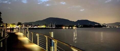 A waterfront promenade at dusk with a calm river, distant mountains, and a well-lit walkway.