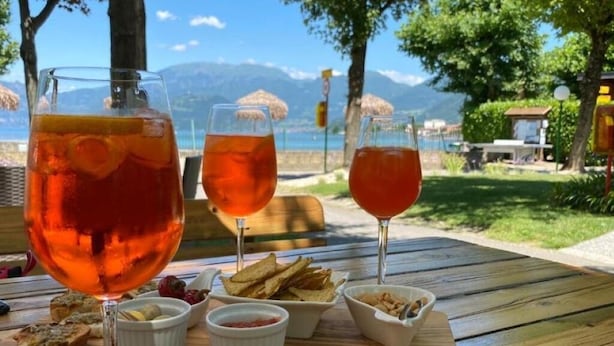 A table with three glasses of red beverage, small bowls of food, and a scenic view of mountains and water. - Duomo Apartment (Iseo)