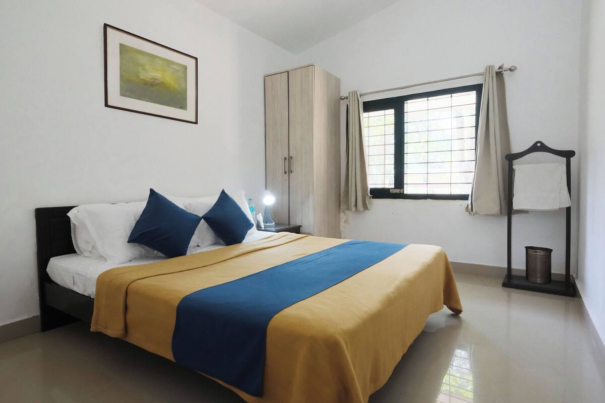 A neatly made bed with a blue and yellow bedspread, a bedside lamp, a wardrobe, a window with curtains, and a framed picture on the wall.