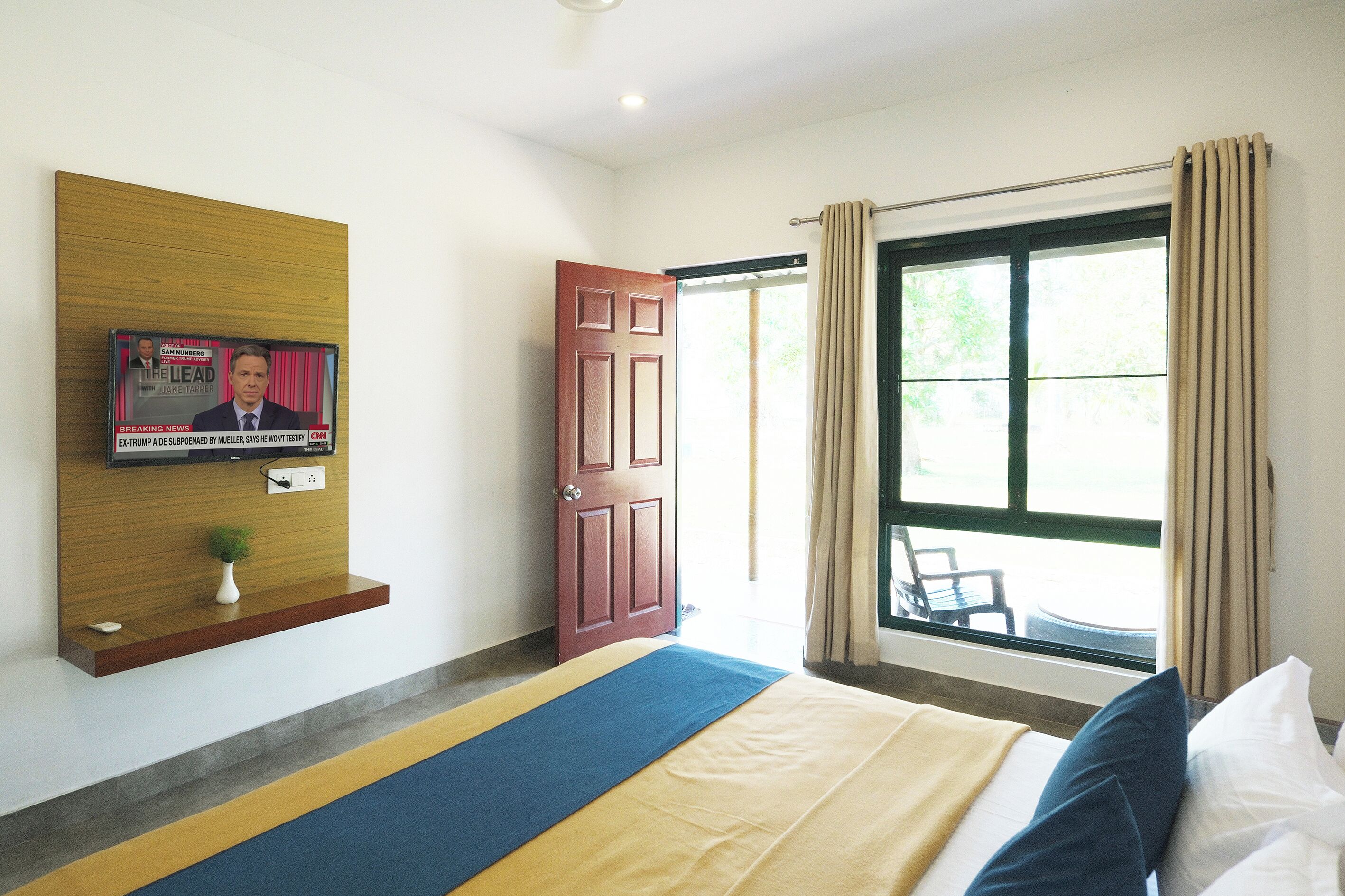A hotel room with a bed, a TV mounted on the wall, a door, and a window with curtains.