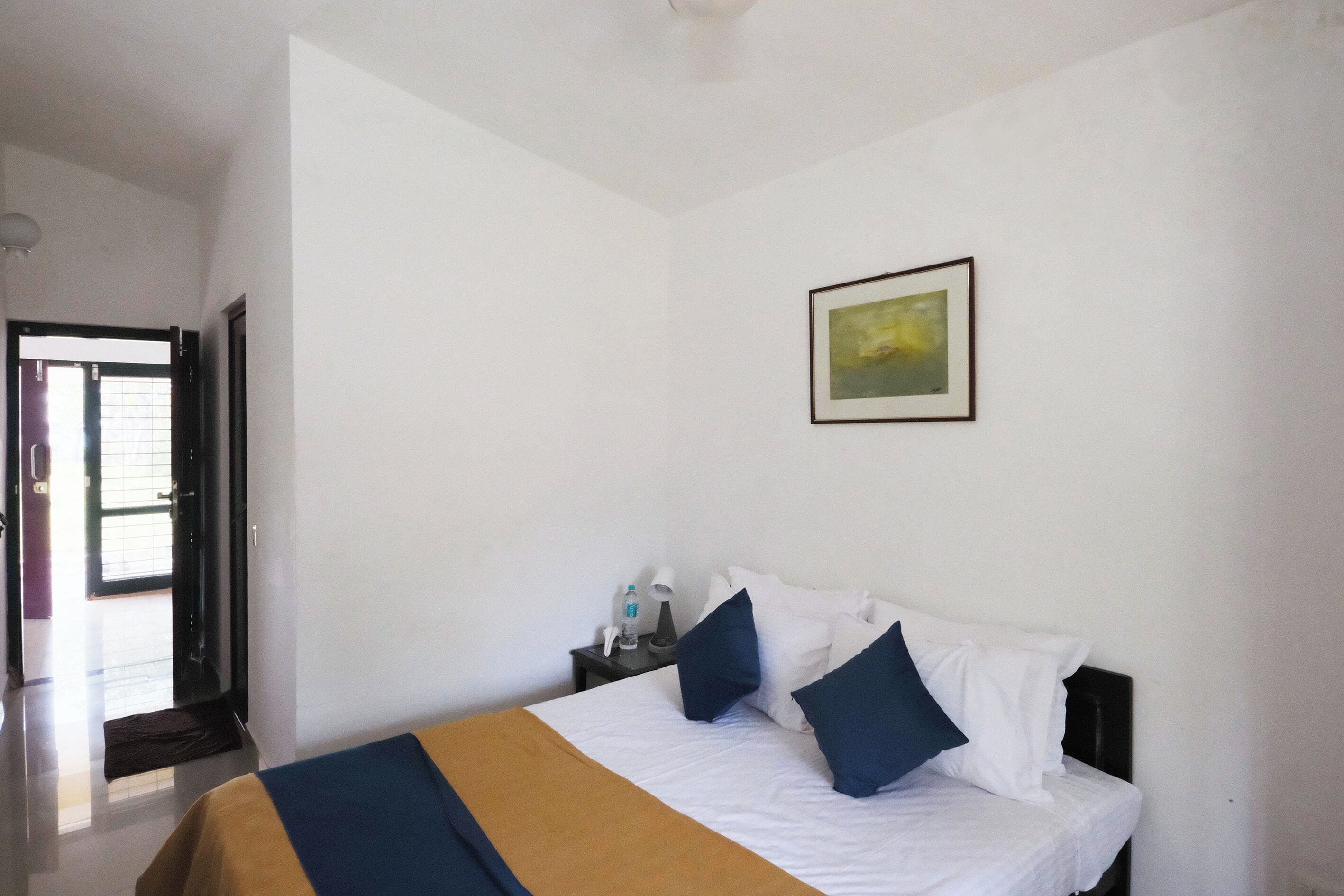 A hotel room with a bed, a nightstand with a lamp, a framed picture on the wall, and a door leading to the outside.